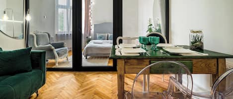 Una sala moderna con piso de madera en patrón espiga, un sofá verde, una mesa de comedor con sillas transparentes y un gran ventanal corredero que da a un dormitorio con cama y ventana.