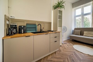 Eine moderne Küche mit Kaffeemaschine, Wasserkocher und Spüle. Auf der Fensterbank steht eine Pflanze, ein Ventilator und im Wohnbereich ein Sofa.
