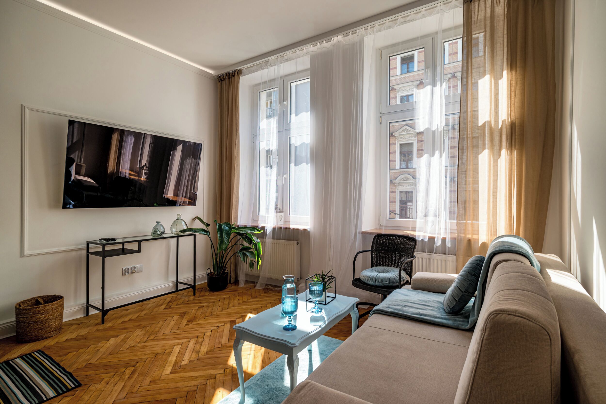 Una sala de estar con un sofá, una mesa de centro, un televisor montado en la pared y un ventanal con cortinas.