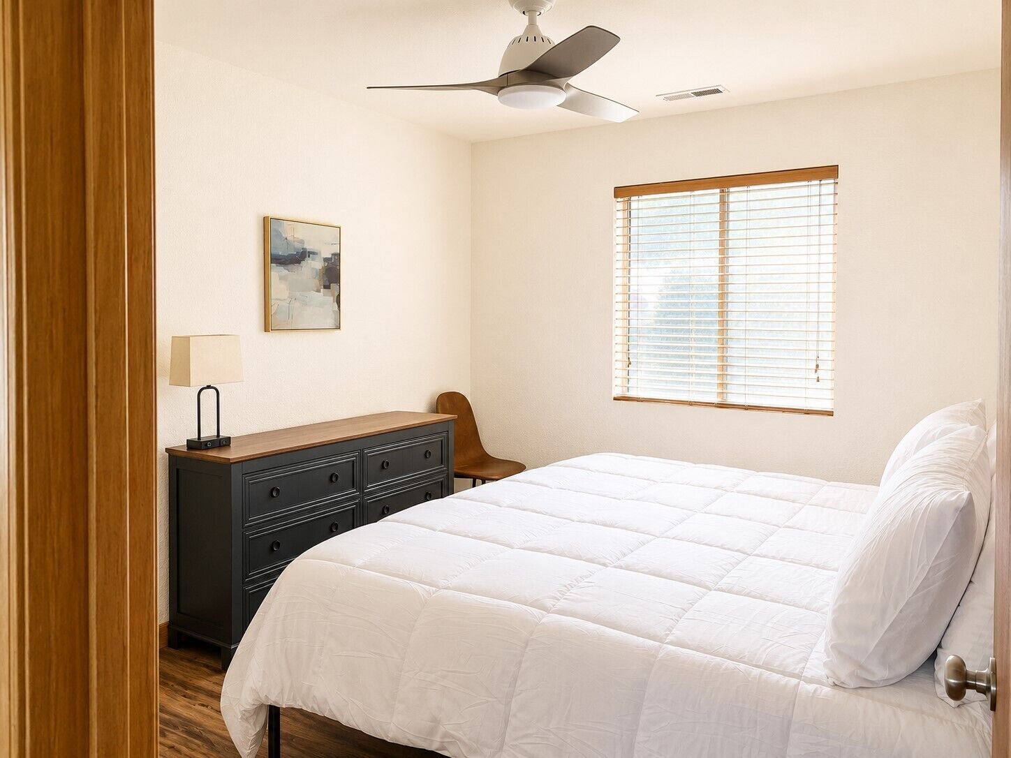 Un dormitorio con cama, cómoda, silla, ventilador de techo y ventana con persianas.
