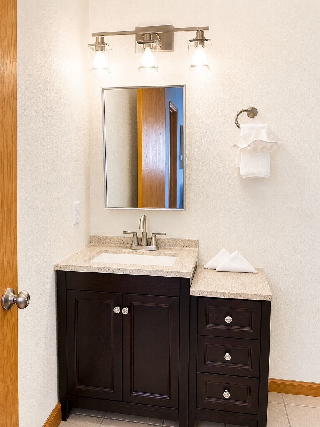 Un baño con tocador de madera oscura, un lavabo blanco y un espejo encima. Hay tres luces empotradas en la pared sobre el espejo.