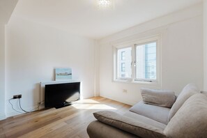 Ein minimalistisch eingerichtetes Wohnzimmer mit Flachbildfernseher, einem Fenster mit Jalousien und einem hellen Sofa.