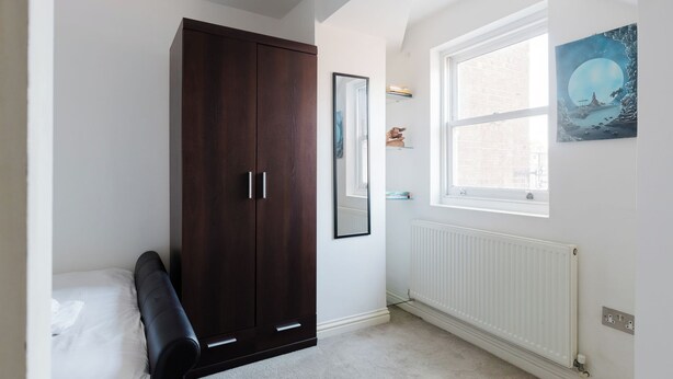 Un dormitorio con un armario de madera oscura, un radiador blanco, una ventana con vistas al exterior y un cuadro en la pared.