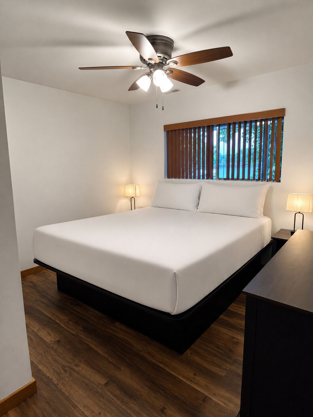 Un dormitorio con cama, ventilador de techo, ventana con persianas y dos lámparas de noche.