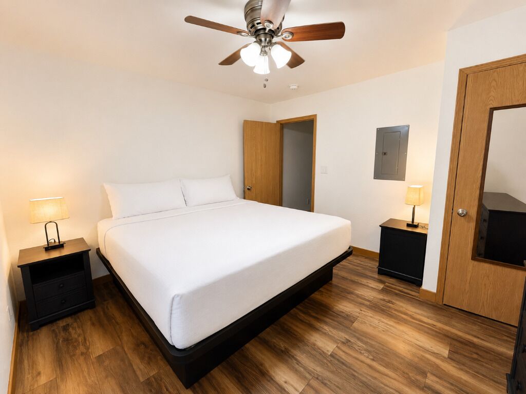 A bedroom with a bed, wooden flooring, a ceiling fan, and two bedside tables with lamps.