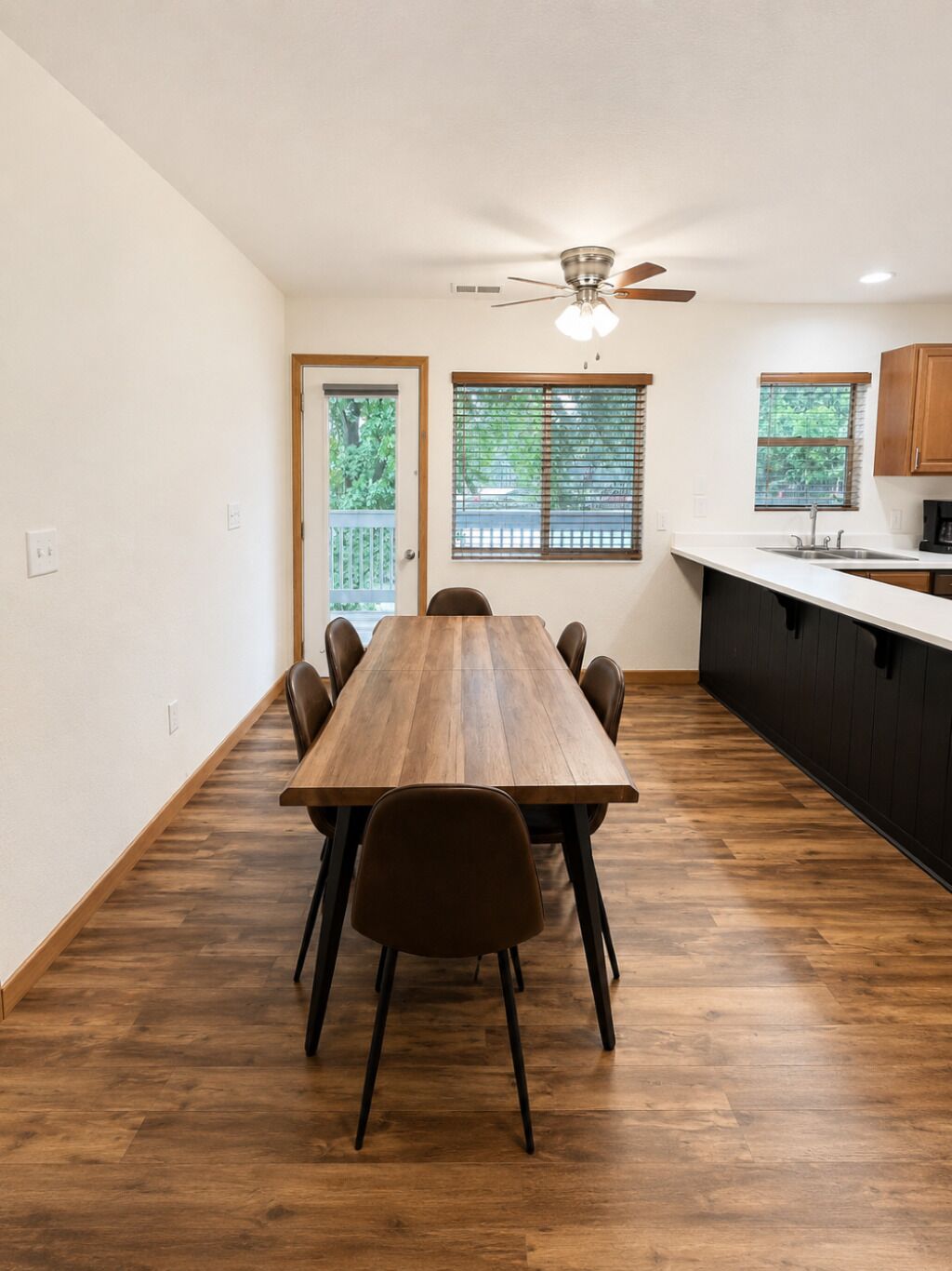 Un comedor con mesa y sillas de madera, un ventilador de techo y una cocina con pileta y alacenas.