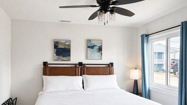 A hotel room with a bed, a ceiling fan, a window with curtains, and artwork on the wall.