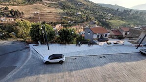 A paved area with a white car parked, surrounded by a low wall and a few trees.