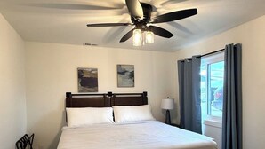 A hotel room with a bed, ceiling fan, artwork on the wall, and a window with curtains.