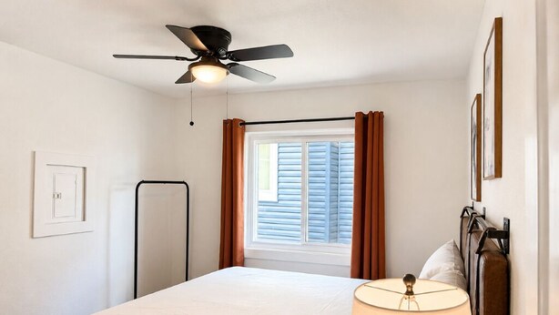 A bedroom with a bed, a ceiling fan, a window with curtains, and a wall-mounted lamp.