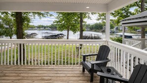A covered outdoor area with a wooden deck, two Adirondack chairs, and a view of a lake with boats.