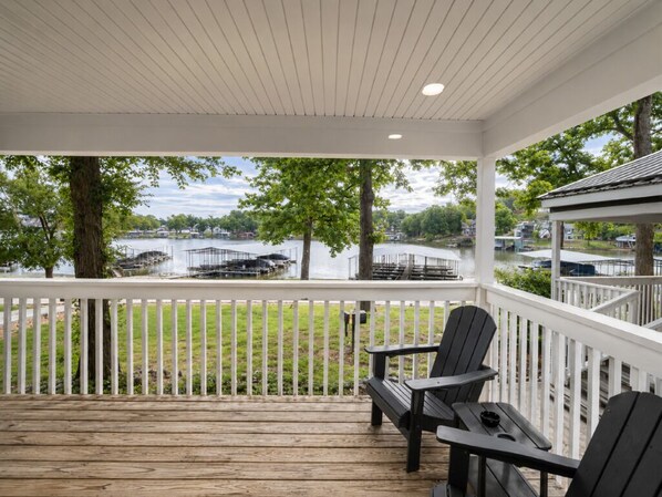 Un espace extérieur abrité, doté d’une terrasse en bois, de deux fauteuils Adirondack et offrant une vue sur un lac où naviguent des bateaux.
