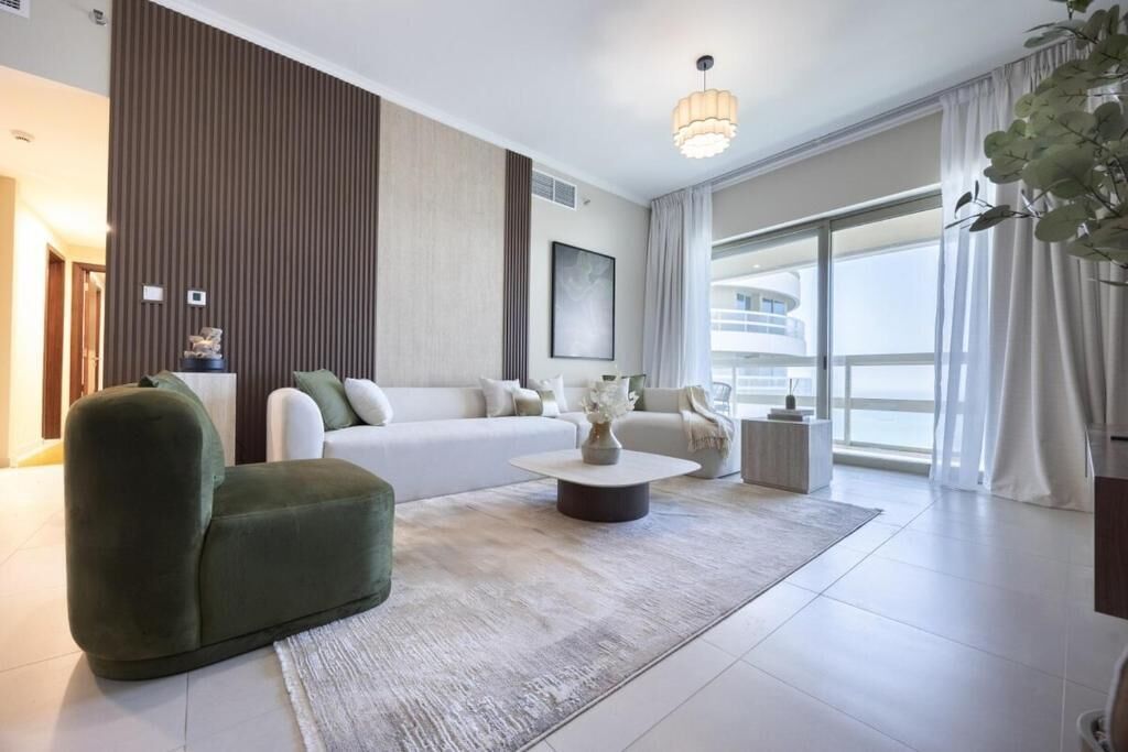 A modern living room with a sofa, a green armchair, a coffee table, and a large window offering a view of a building.
