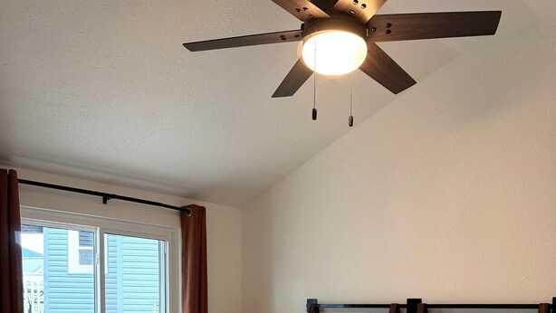 A bedroom with a bed, a ceiling fan, a window with curtains, and a lamp.
