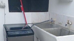 A utility room with a trash bin, a mop, a blue bucket, and a window with a black screen. - Cozy 2-bedroom apartment located just 5 minutes from Nadi International Airport. (Martinar)