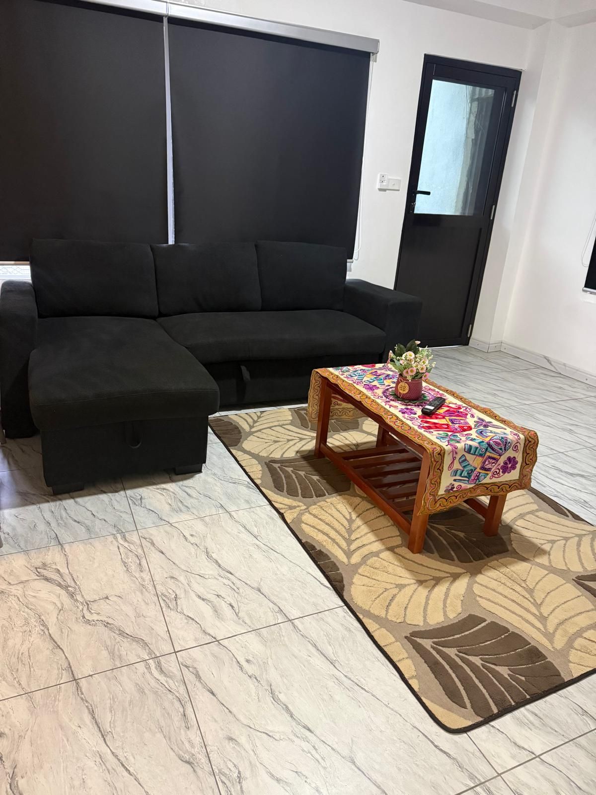 Un salon avec un canapé noir, une table basse en bois recouverte d’une nappe colorée et un tapis à motifs.