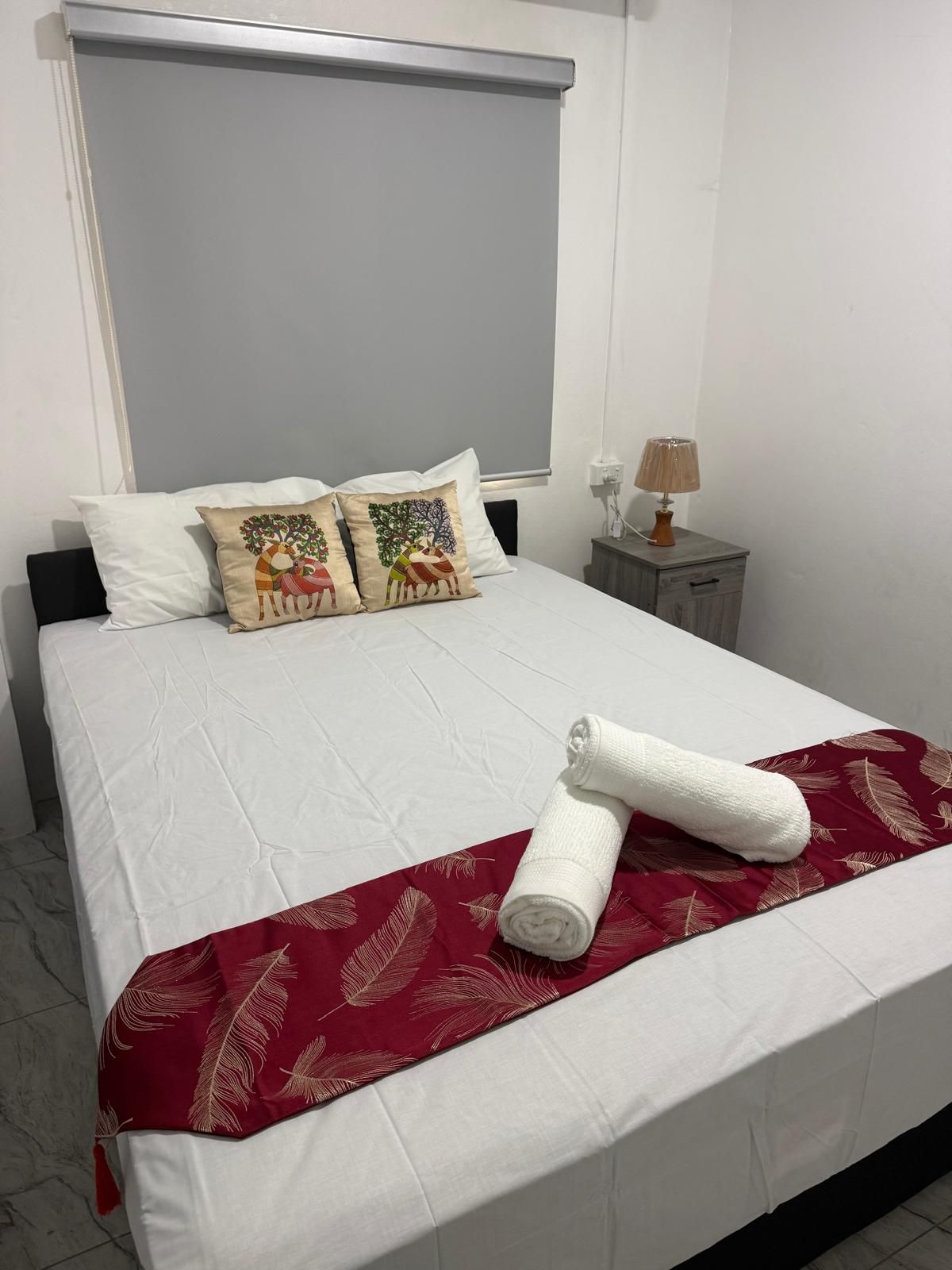 Een netjes opgemaakt bed met witte lakens, twee decoratieve kussens, een rode verenplaag en een opgerolde handdoek.