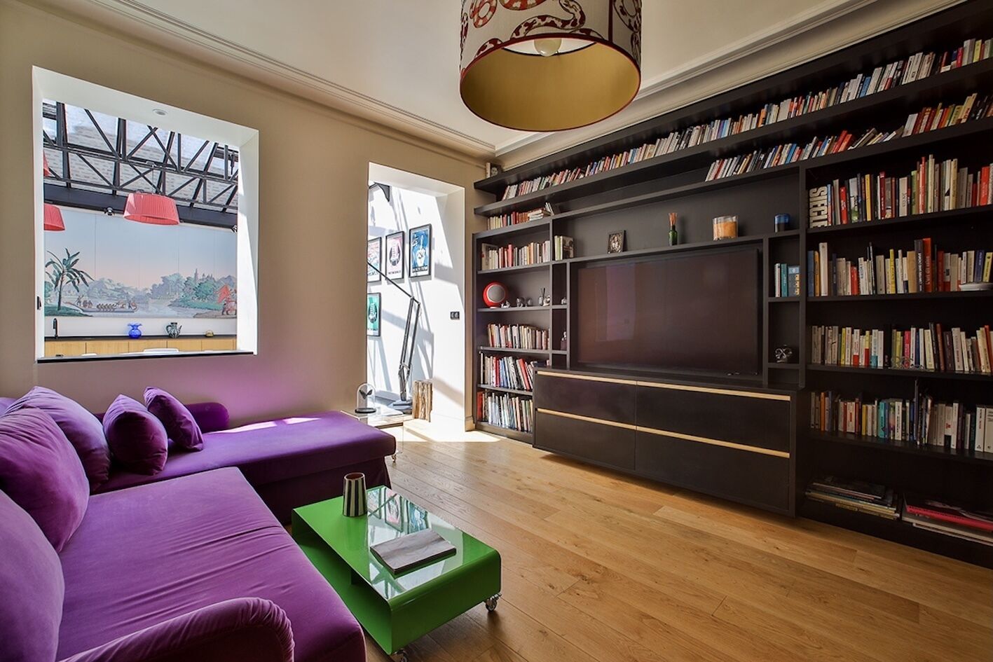 A modern living room with a purple sofa, a green coffee table, a large bookshelf, and a television.