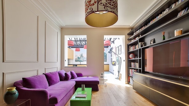 A modern living room with a purple sectional sofa, a green coffee table, and a bookshelf filled with books. - Villa Amore (Paris)