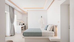 A modern bedroom with a built-in bed, bedside tables, a hanging robe, and a bathroom with a sink and mirror. - Parolea Luxury Living Hotel (Paros)