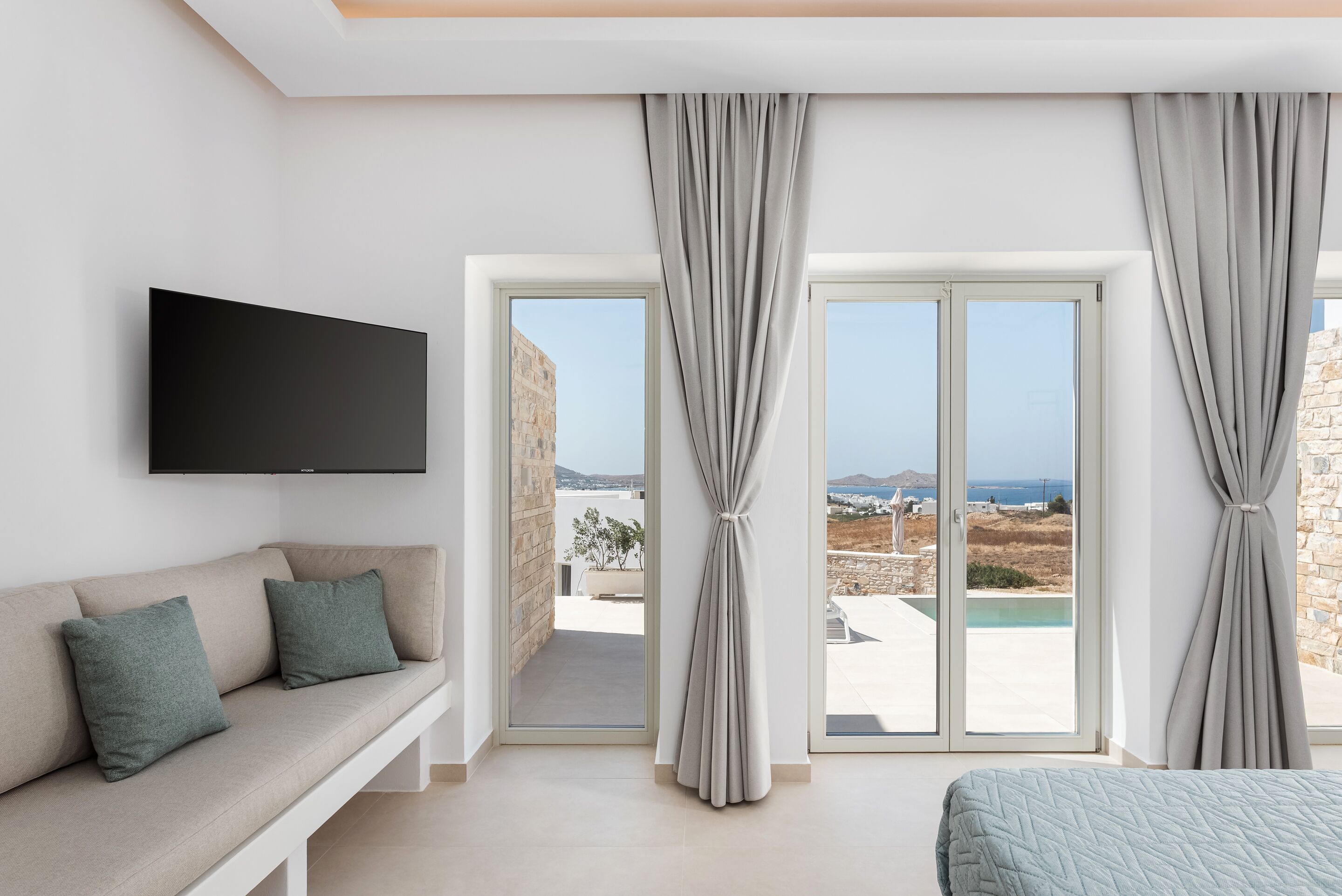 A modern hotel room with a sofa, a bed, a TV, and a view of the sea through large windows.