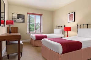 Quarto de hotel com duas camas, uma escrivaninha e uma cadeira. Há duas luminárias vermelhas, uma janela com persianas e duas quadros nas paredes.