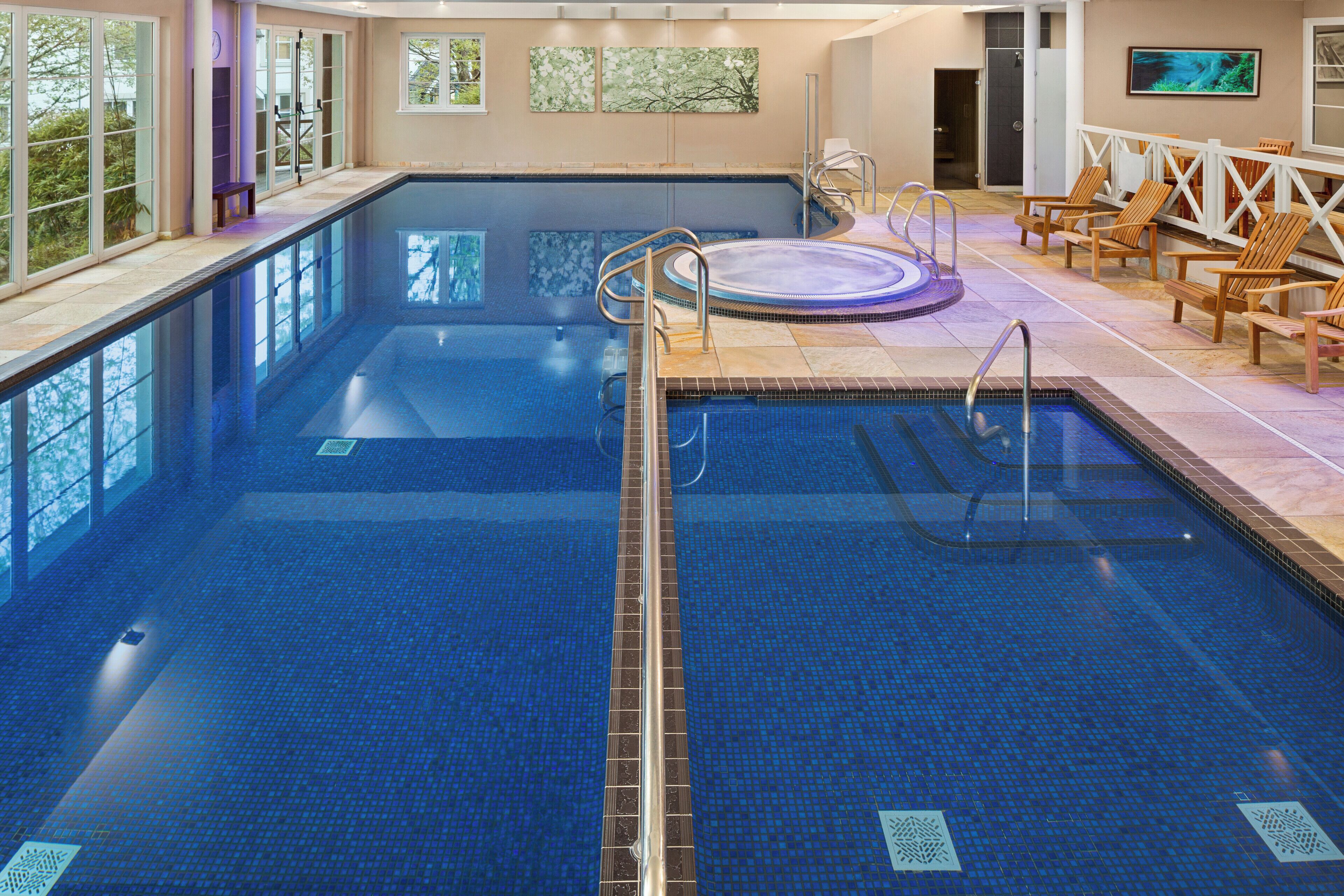 A large indoor swimming pool with a hot tub, surrounded by wooden deck chairs and a glass-enclosed area with windows.