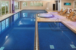 Una amplia piscina interior con un jacuzzi, rodeada de sillones de madera y una zona cubierta con ventanas.