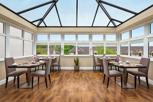 Un ambiente luminoso con varias mesas y sillas, amplias ventanas y vista a casas.