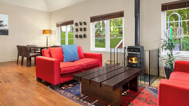 A cozy living room with a red sofa, a coffee table, and a fireplace. There is a dining area with a table and chairs, and a window with a view of greenery. - Duchally Hotel & Country Estate (Auchterarder)