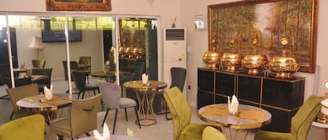A room with round tables, green chairs, a fireplace with golden urns, and a large painting on the wall.
