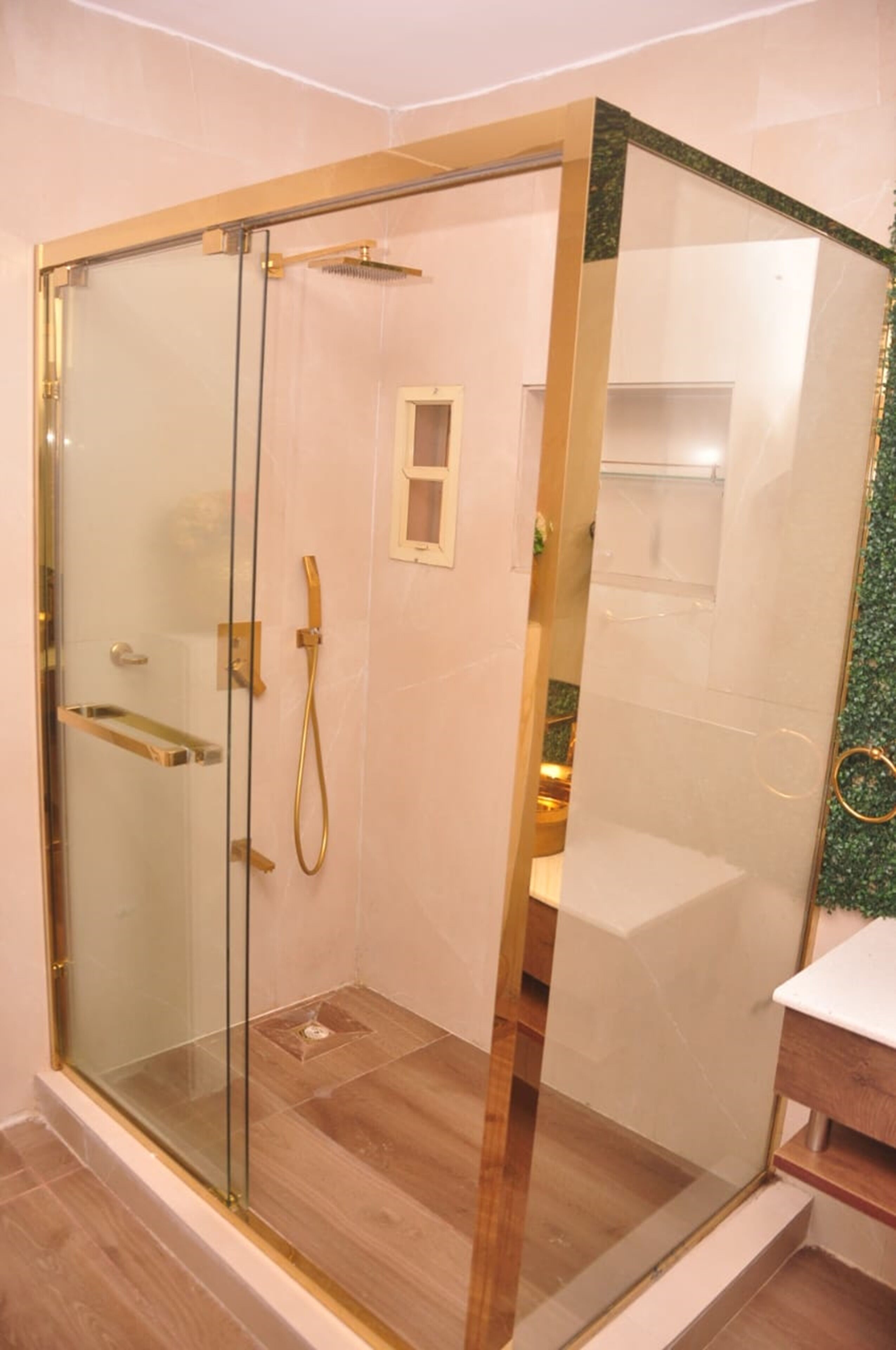 Une douche fermée par des parois de verre, avec un cadre doré, une douchette à main et une petite fenêtre.