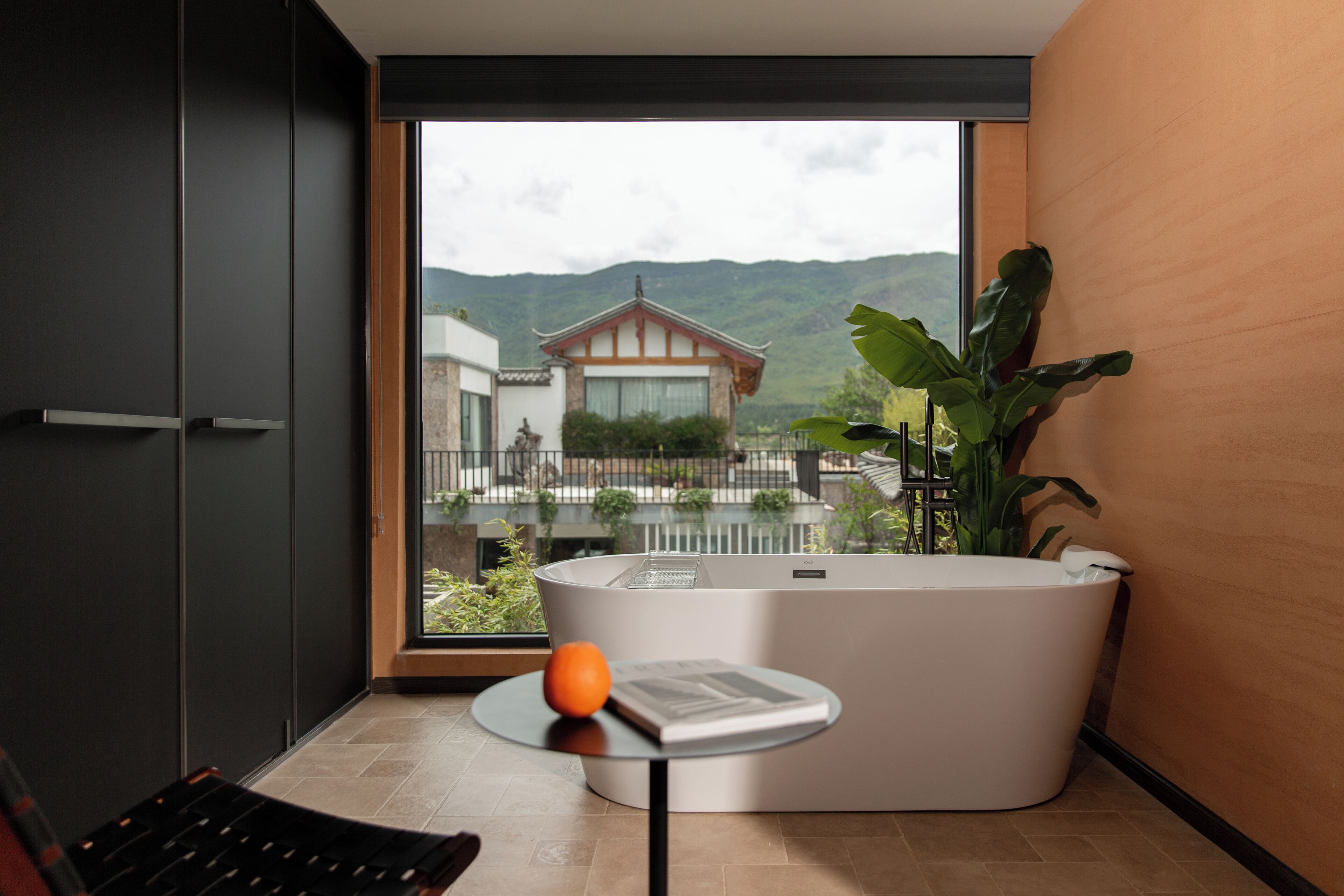 A modern bathroom with a large bathtub, a small table with a book and an orange, and a view of a traditional house and mountains through a large window.