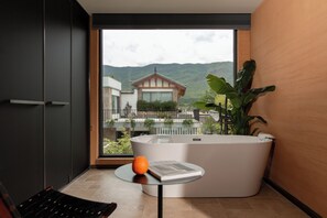 Un baño moderno con una bañera grande, una mesita con un libro y una naranja, y vistas a una casa tradicional y montañas a través de un ventanal amplio.
