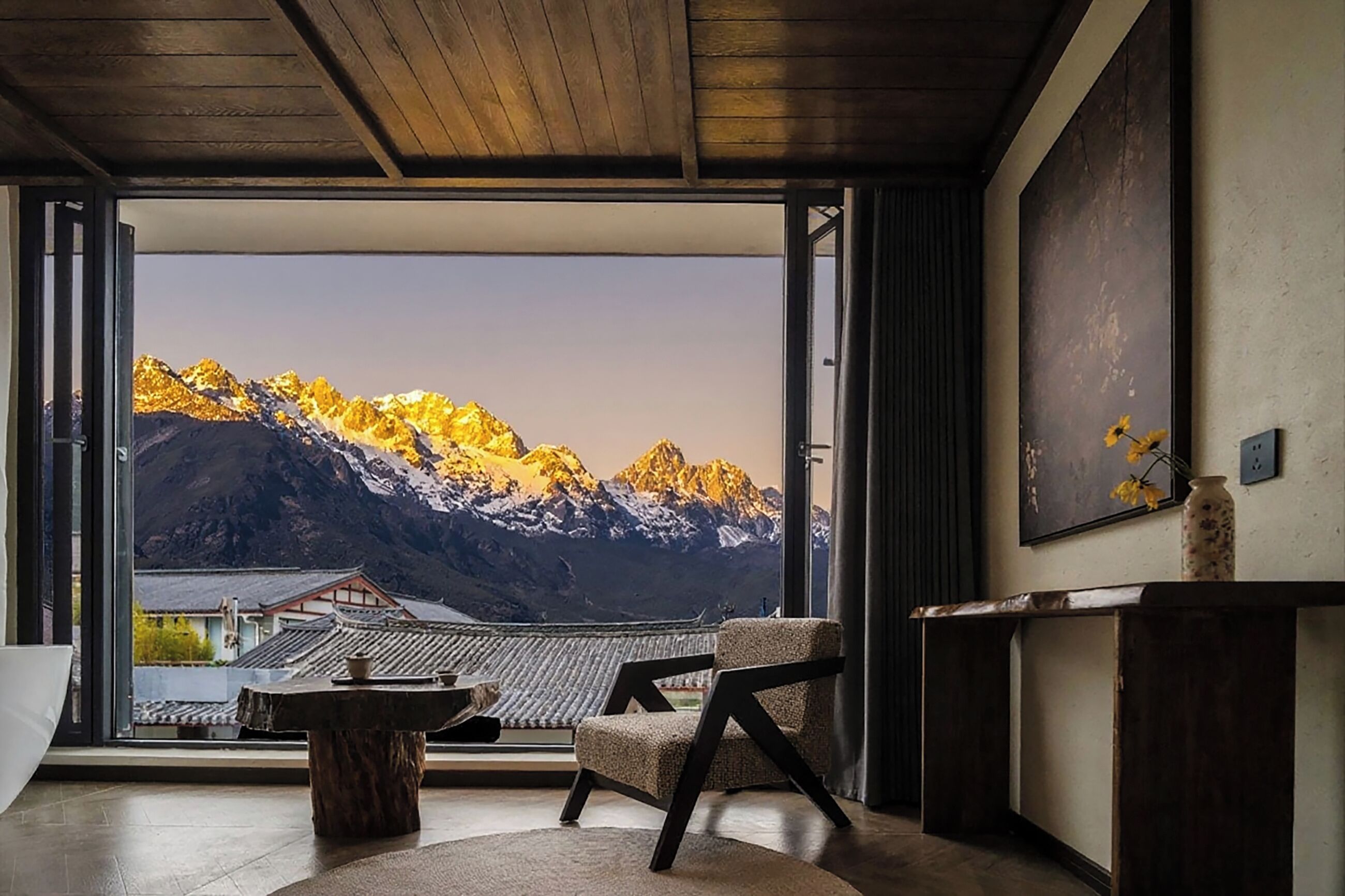 Habitación con una ventana grande que ofrece vistas a la montaña, una silla tradicional, una mesita y un jarrón con flores.