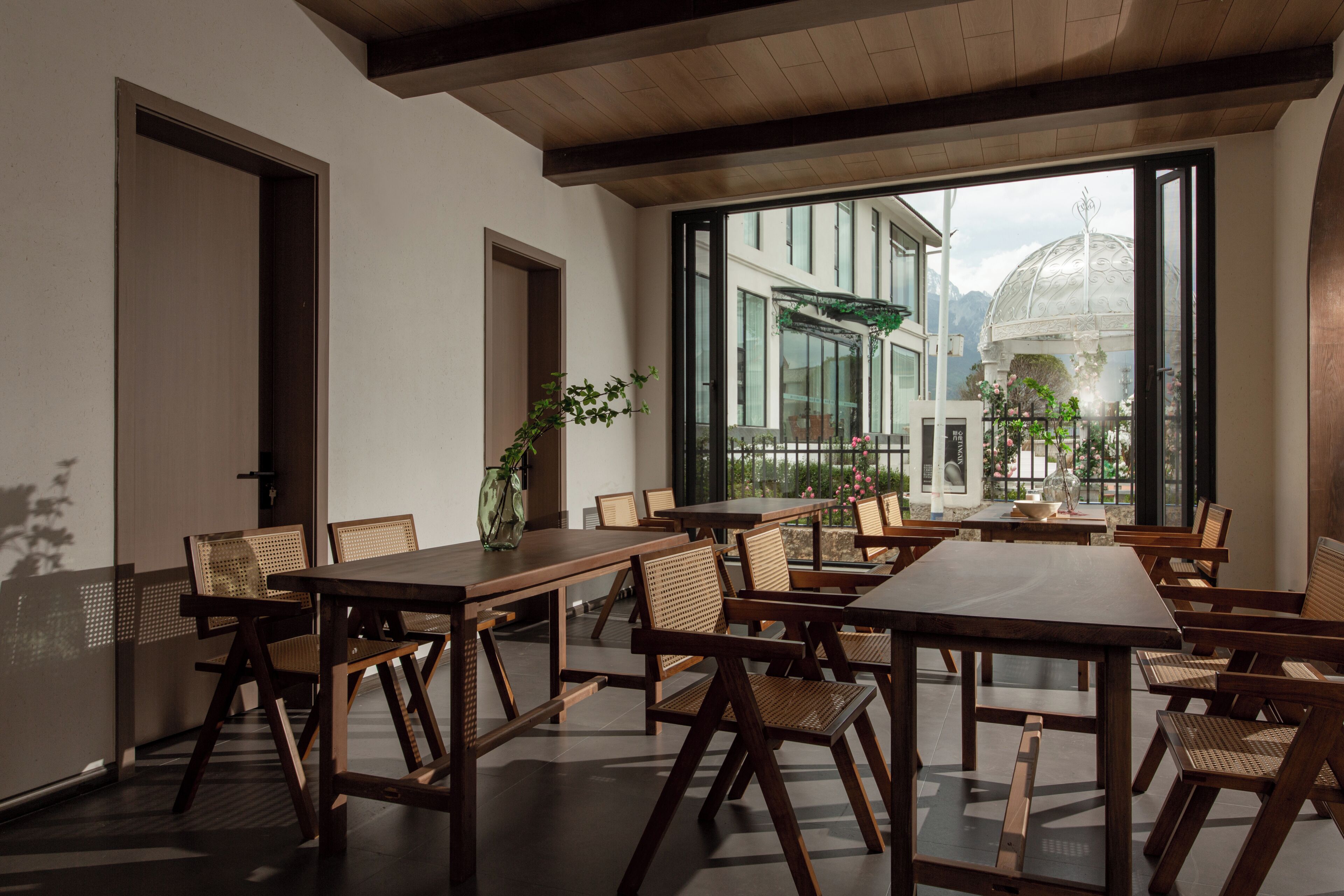 Une salle à manger spacieuse, dotée de tables et de chaises en bois, de grandes fenêtres offrant une vue sur un jardin et un bâtiment surmonté d’une coupole.