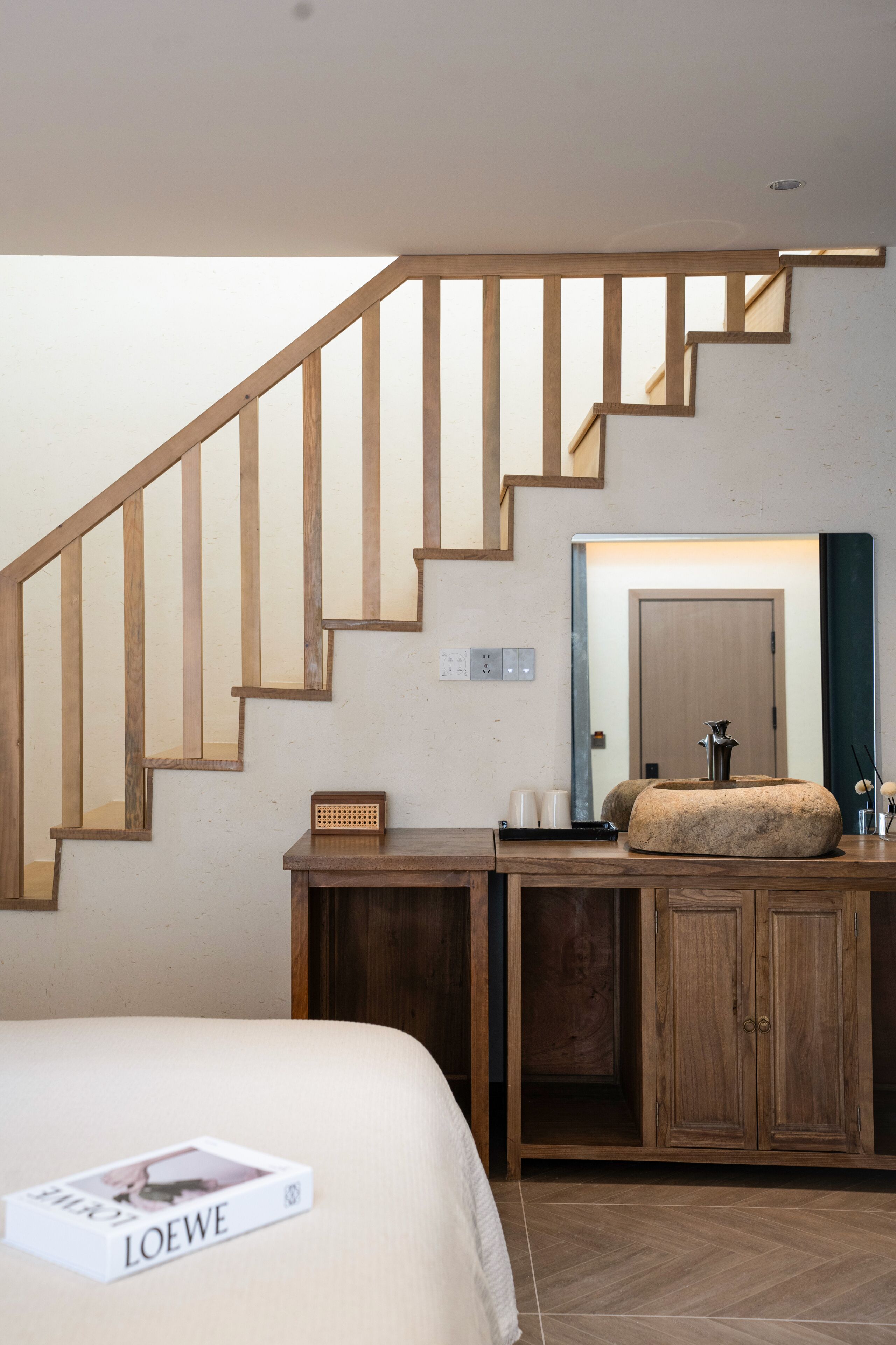 A bedroom with a wooden staircase, a bed with a magazine, a desk with a mirror, and a stone basin.