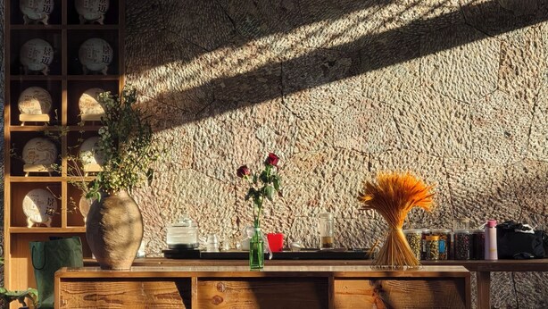 A wooden reception desk with a textured stone wall in the background, a vase with flowers, and a small table with various items.