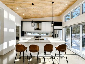 Een moderne keuken met een centraal eiland, hangende verlichting en uitzicht op een besneeerd landschap door grote ramen.
