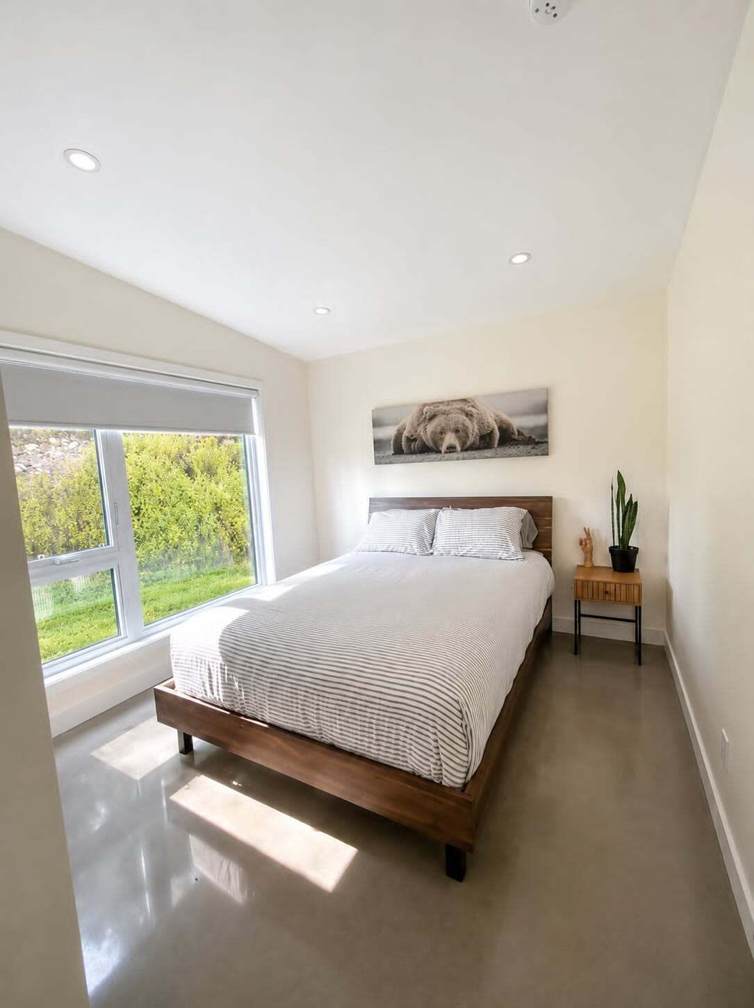 Un dormitorio moderno con un ventanal grande, una cama con ropa de cama a rayas, una obra de arte con temática de oso sobre la cama y una mesita de noche pequeña con una planta.