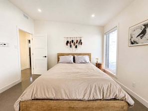 A bedroom with a large bed, a wooden headboard, a wall-mounted macrame decoration, a framed picture, and a window with a view.