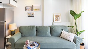 Ein modernes Wohnzimmer mit einem grauen Sofa, einem kleinen Couchtisch mit Zeitschriften, einer Lampe und gerahmten Kunstwerken an der Wand.