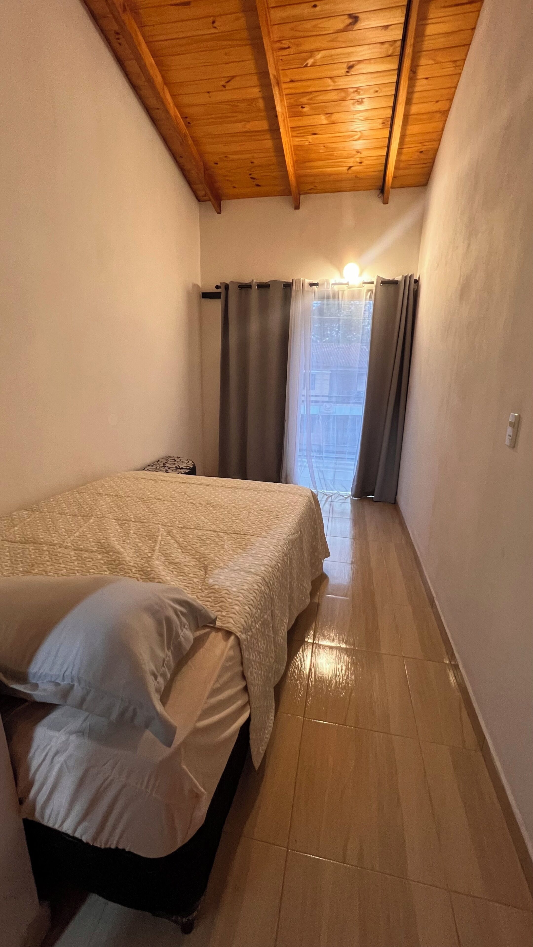 Una habitación pequeña con cama, techo de madera y vista por una ventana con cortinas.
