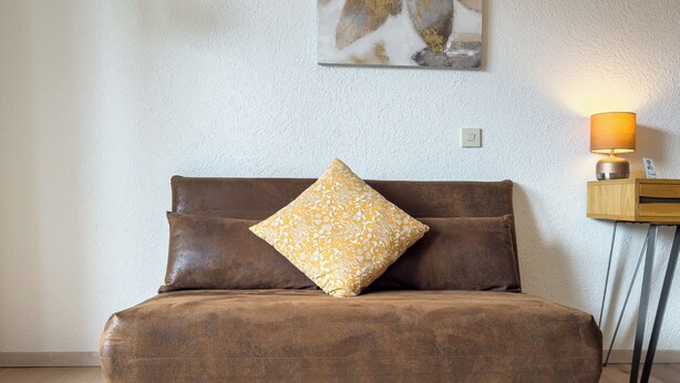 Ein braunes Sofa mit einem gemusterten Kissen, eine strukturierte Wand, ein kleiner Holztisch mit einer Lampe und ein Gemälde über dem Sofa.