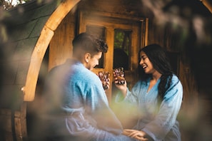 Twee personen zitten aan een houten tafel met drankjes, in een rustieke omgeving met een houten deur en boogvormige doorgang.