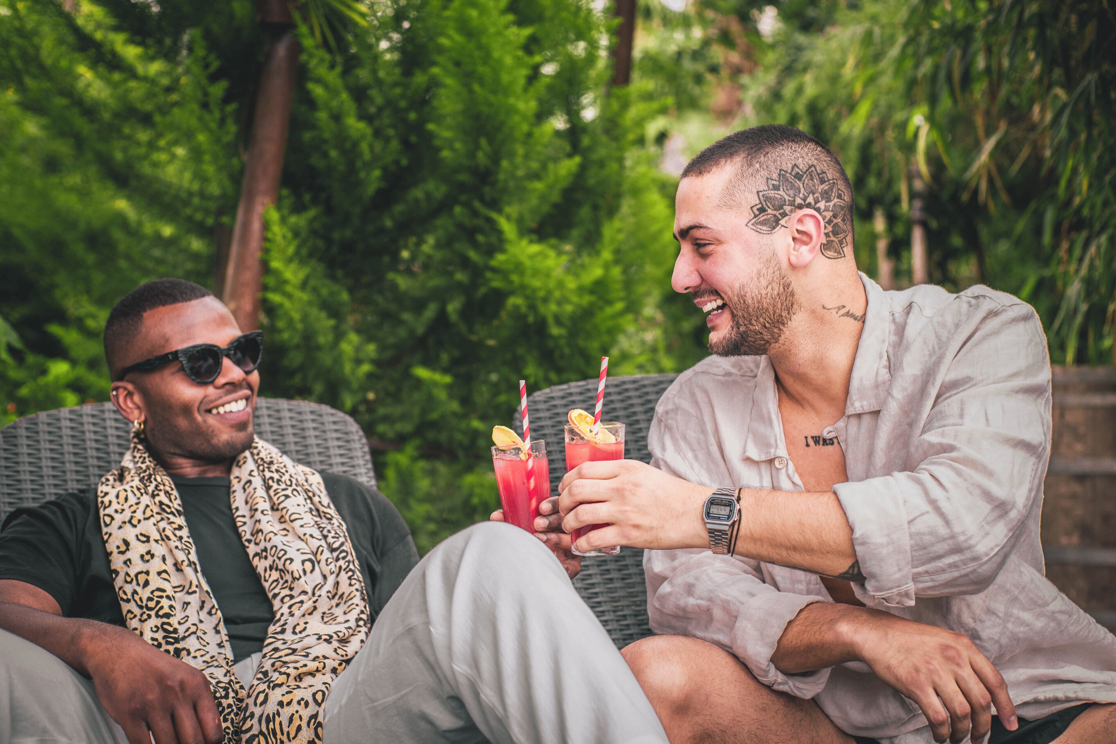 Two individuals seated on a wicker sofa outdoors with greenery in the background.