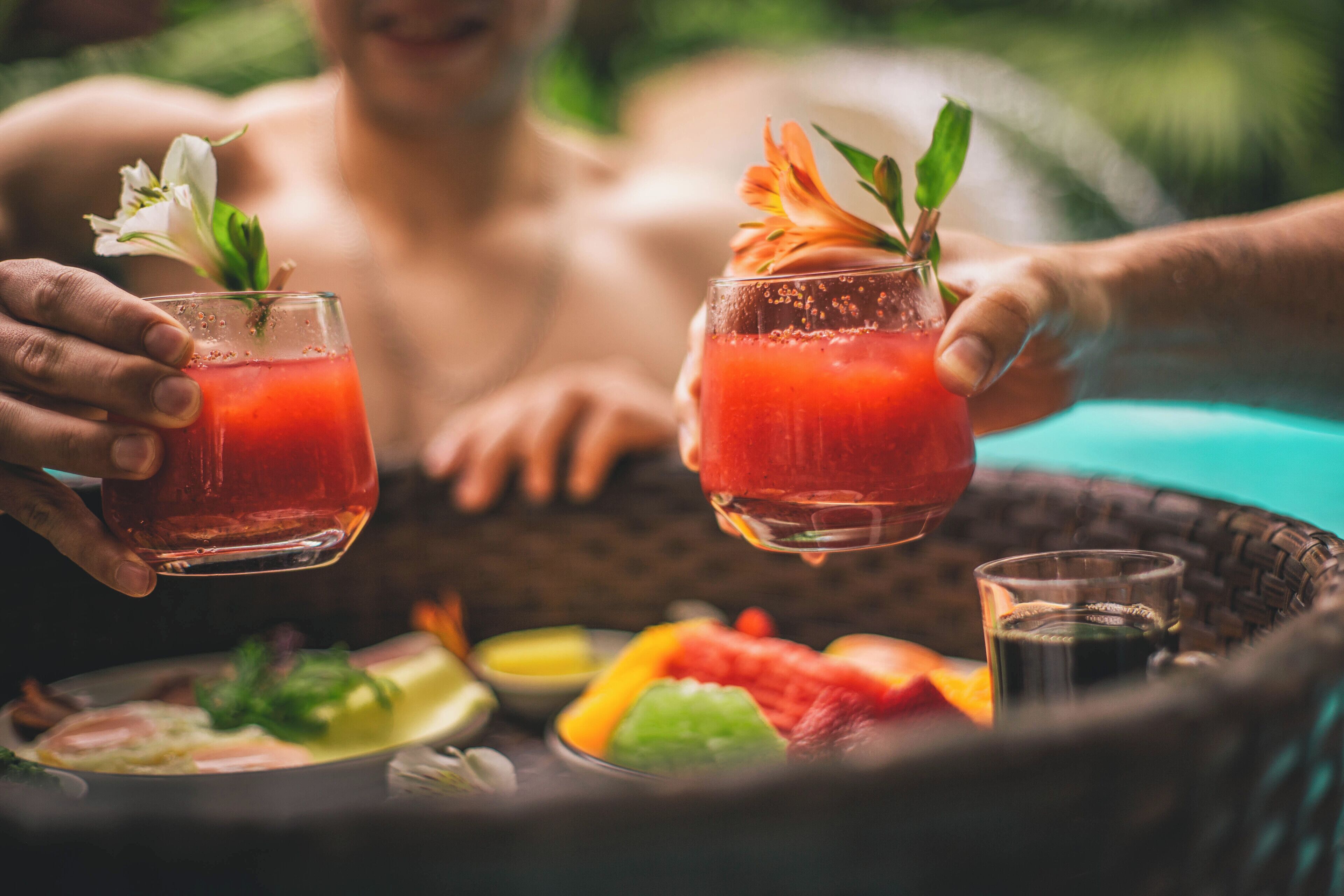Deux personnes tenant des verres contenant une boisson rouge, avec un panier de fruits et un petit verre contenant un liquide foncé sur une table.