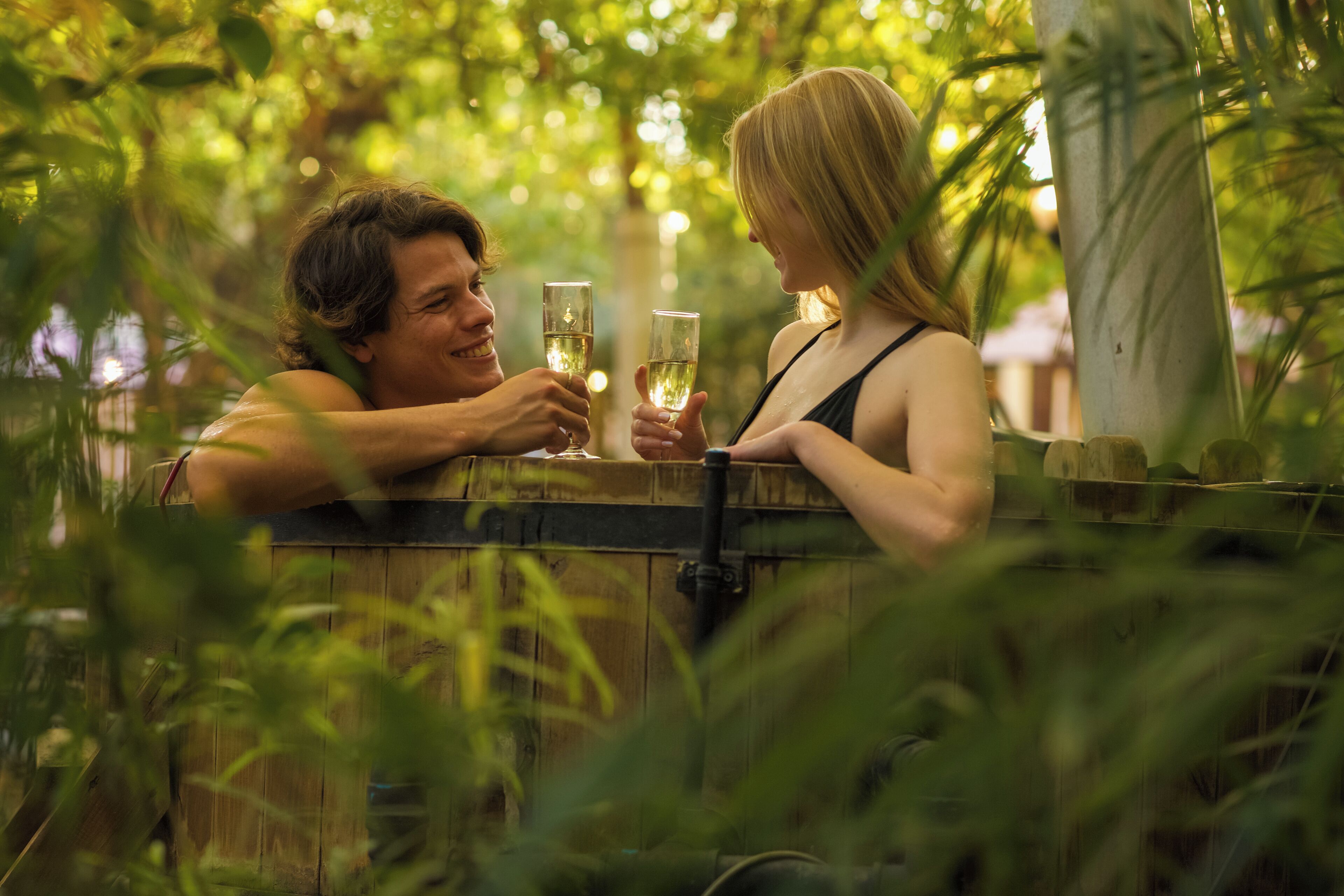 A couple sitting at a wooden table outdoors, holding glasses of a clear liquid, likely wine, with greenery in the background.