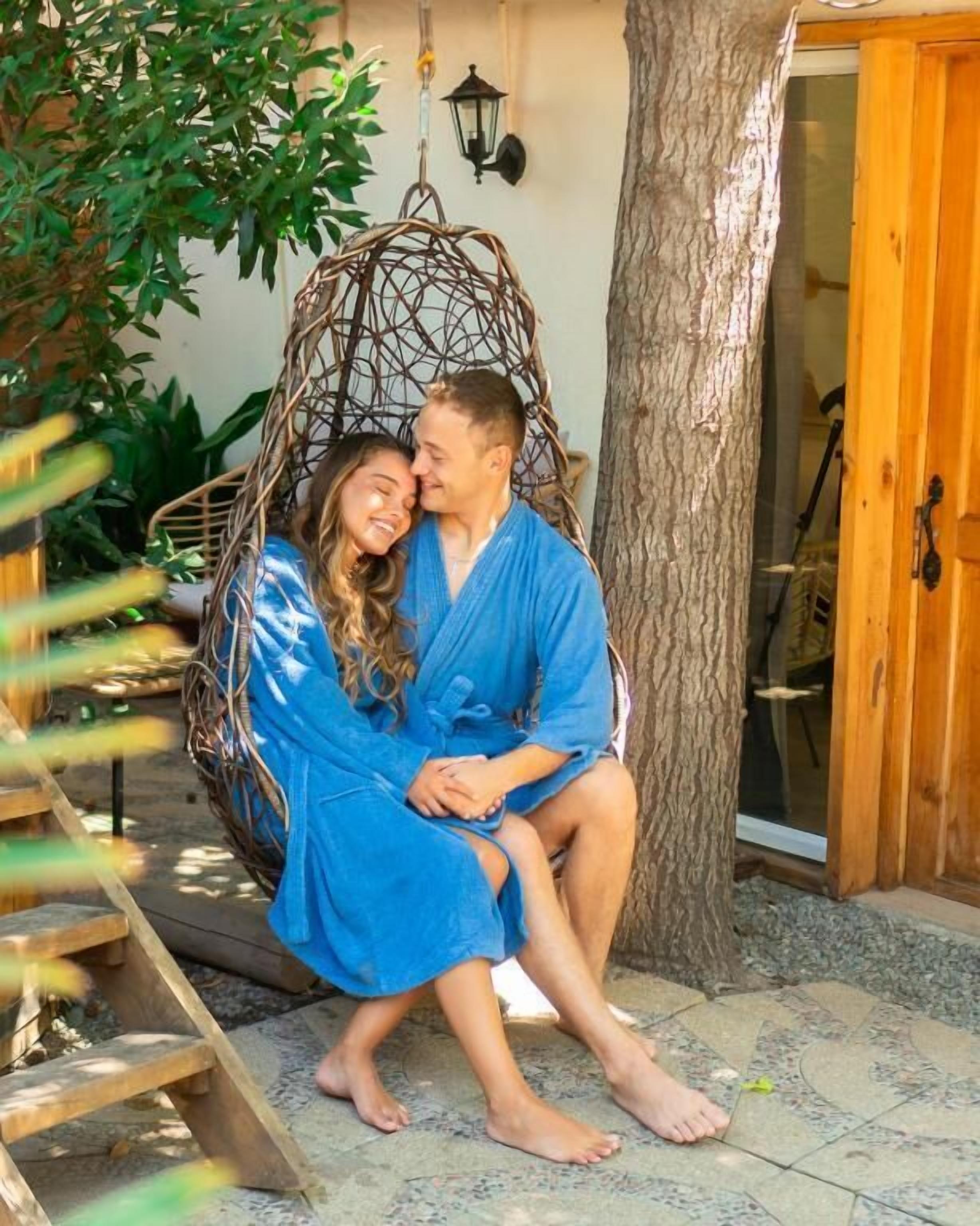 A couple sitting in a wicker swing chair outdoors, with a tree and a wooden door in the background.