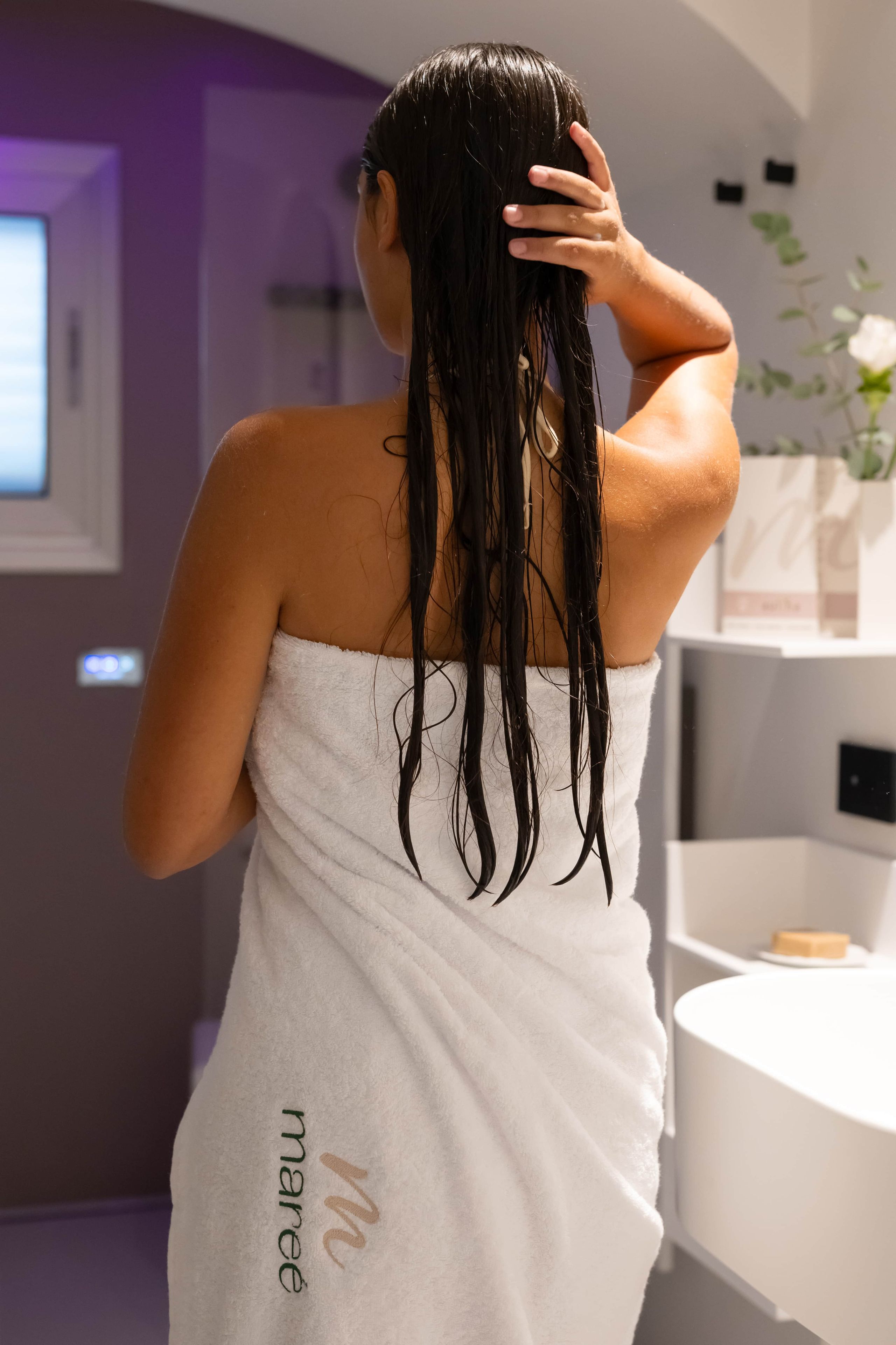 Une personne aux cheveux mouillés, enveloppée dans une serviette, se tient dans une salle de bain qui comprend un lavabo blanc et un miroir.